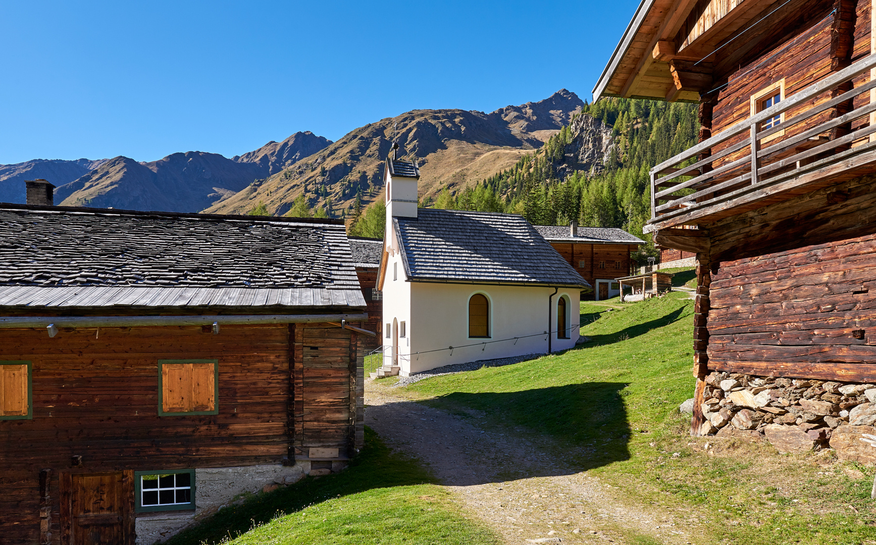 Blick durch die hölzernen Almhütten auf die Schutzengelkapelle auf der Oberstalleralm. Die Häuser...