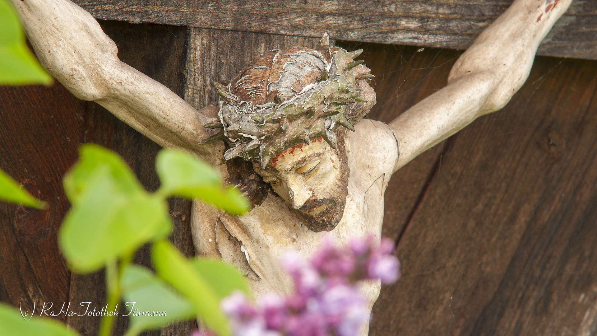 Christus am Wegkreuz mit blühendem Flieder im Vordergrund