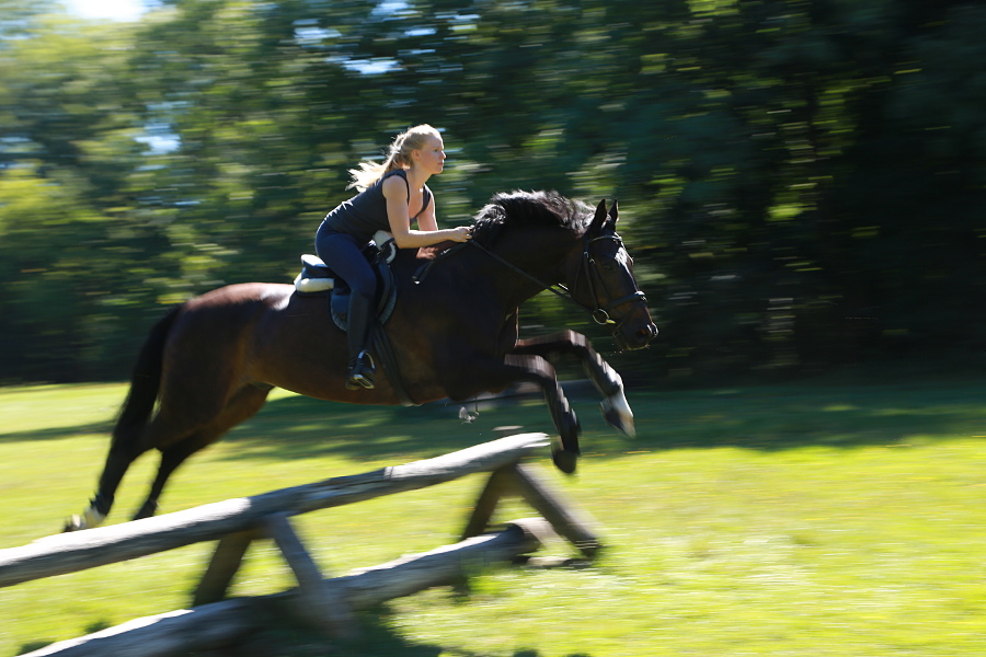 Girl With Golden Hair Rides Black Beauty Over Bar While Photographer Is Moving His Camera