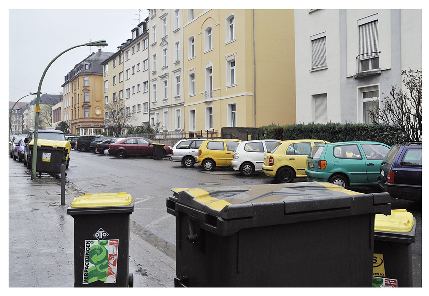 muelltonnen in strasse mit parkenden autos, bogenleuchten und wohnhaeusern.