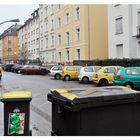 muelltonnen in strasse mit parkenden autos, bogenleuchten und wohnhaeusern.